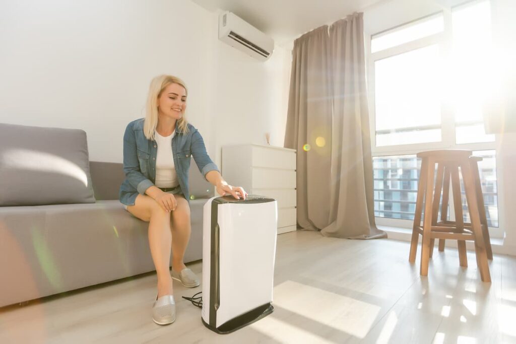 A woman sitting on a sofa in her apartment turning on an indoor air conditioning unit.