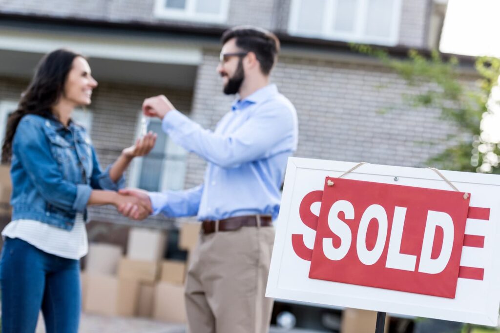 A woman takes the keys from a man after buying a house in front of a large for sale/sold sign