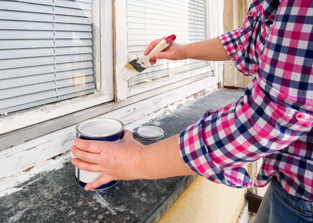 A man paints an exterior window to finish his checklist of late summer home improvement projects.