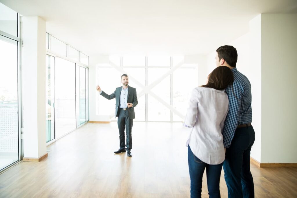 A man in a suit conduction a property showing with a man and a woman in an apartment with large windows