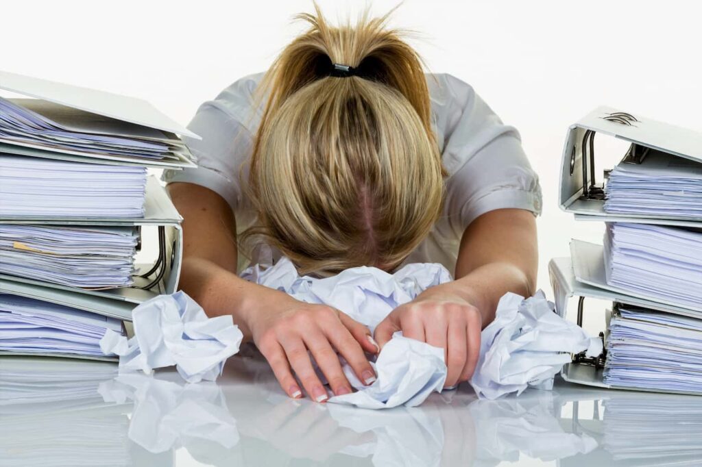 a woman experiencing burnout buries her head in a pile of crumpled papers between two large stacks of binders.