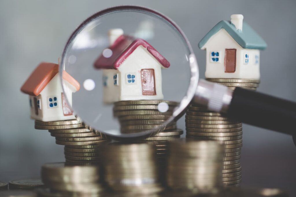 A row of small houses sit on top of piles of coins with a magnifying glass over one to signify a move-out inspection.