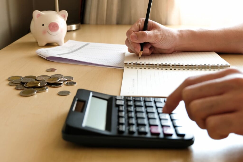 a person sitting at a desk using a pen, pencil, and calculator to add up their property management costs. A piggy bank and coins also sit on the desk.