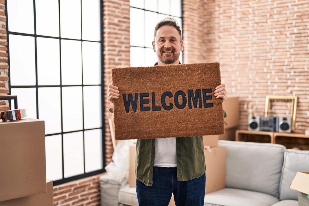 a person holding up a welcome mat in brick-walled room with large windows, symbolizing how an onboarding practice can make new tenants feel welcome in their home.